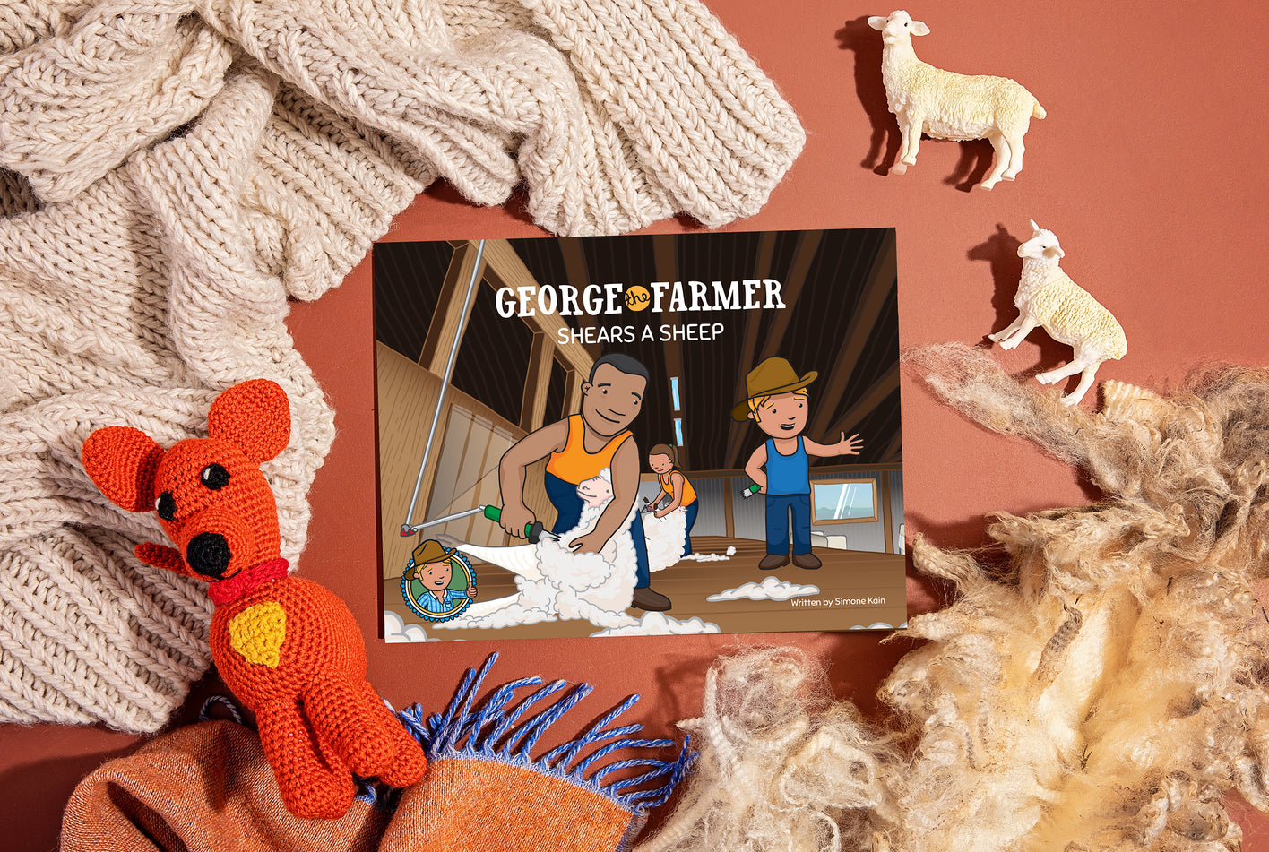 A promotional image featuring a picture book titled 'George the Farmer Shears a Sheep', with illustrations of characters and sheep. There are also small plush toys of a sheep and a farm animal visible.